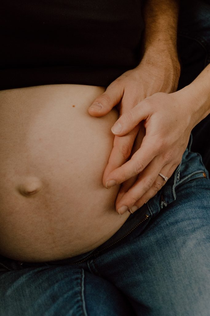 Ventre de femme enceinte en séance maternité lifestyle à domicile, mains posées délicatement sur le ventre en lumière douce, photo professionnelle réalisée par une photographe maternité au Québec.