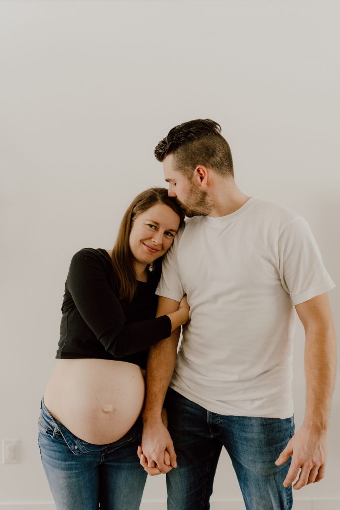 Séance photo maternité lifestyle à domicile, couple debout main dans la main mettant en valeur le ventre de grossesse, photo professionnelle réalisée par une photographe lifestyle au Québec.
