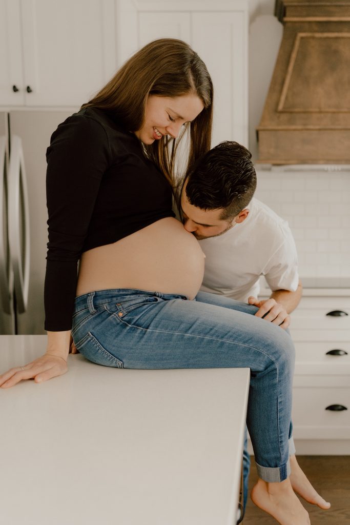 Couple en séance photo maternité lifestyle à la maison, futur papa embrassant le ventre de la maman dans une cuisine lumineuse, photo professionnelle réalisée par une photographe lifestyle au Québec.