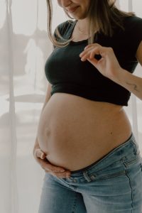Femme enceinte portant un haut noir et un jean, tenant son ventre avec tendresse devant une fenêtre lumineuse