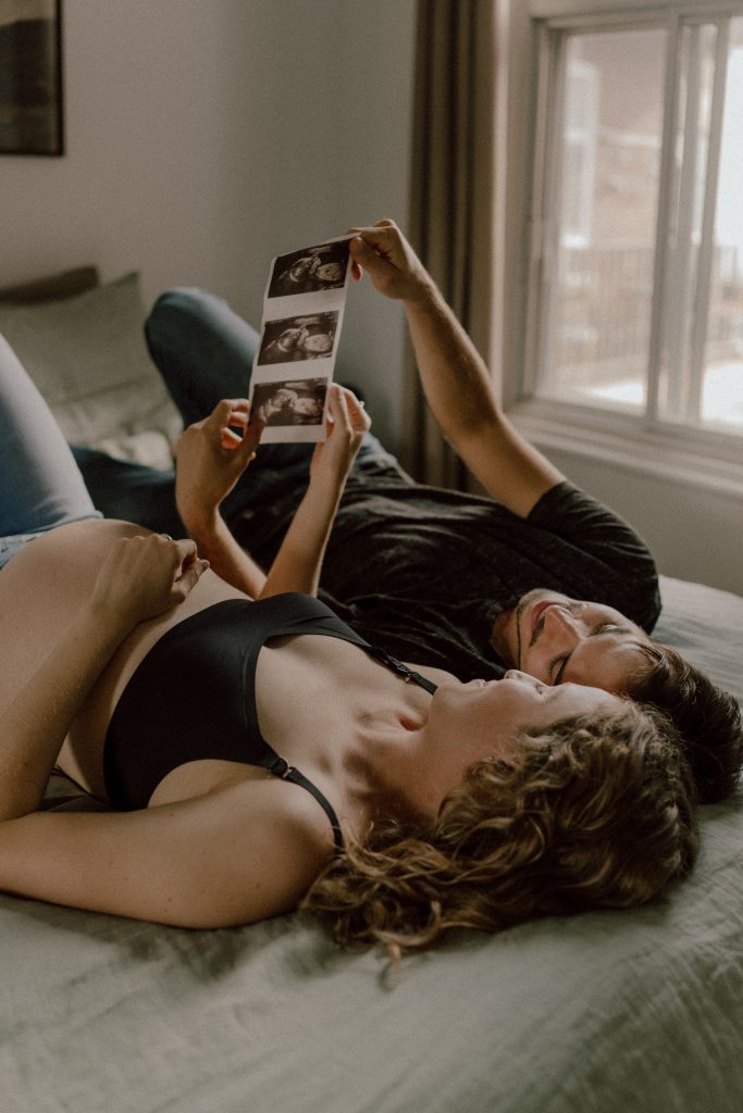 Couple allongé sur un lit, tenant et regardant des échographies de leur futur bébé, créant un moment intime et tendre.