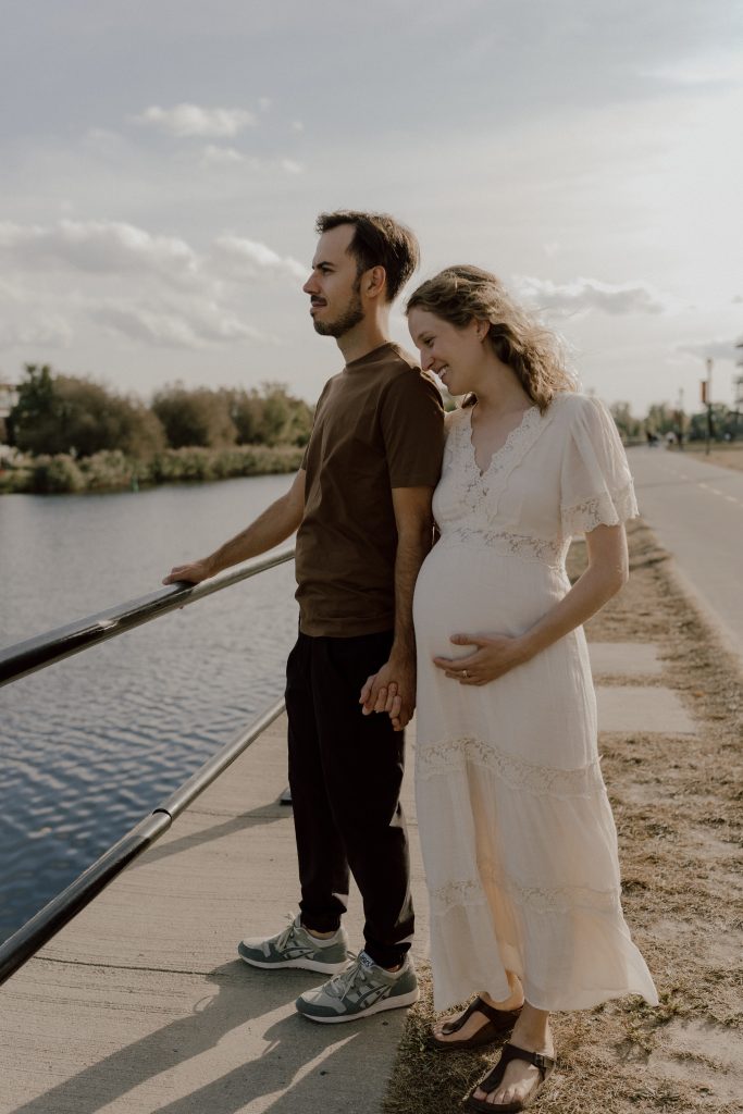 Couple tenant la main, debout près de l'eau lors d'une promenade, la future maman tenant son ventre avec tendresse, dans un moment de complicité et de sérénité.