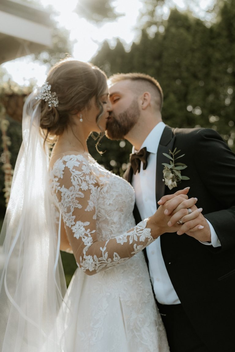 Mariée portant une robe en dentelle et voile embrassant le marié en smoking, dans un décor naturel avec une lumière douce et romantique.