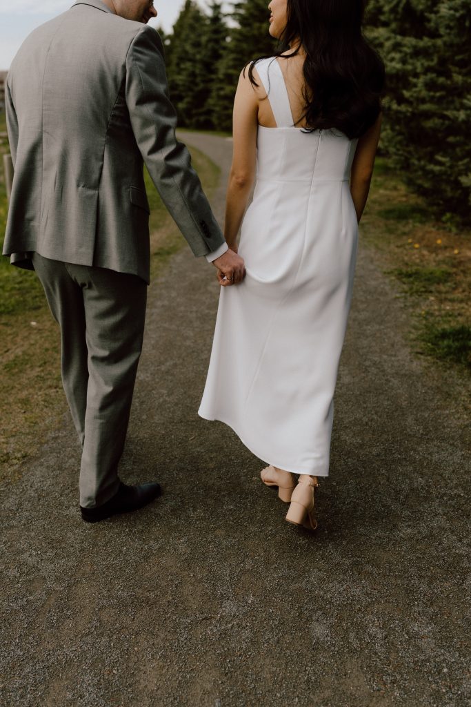 Couple de mariés marchant sur un chemin bordé de verdure, le marié tenant délicatement la main de la mariée vêtue d'une robe blanche simple et élégante.