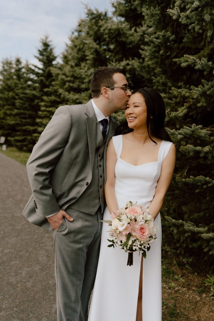 Moment tendre entre les mariés, le marié embrassant la mariée sur la tête alors qu'elle tient son bouquet dans un cadre naturel et chaleureux.
