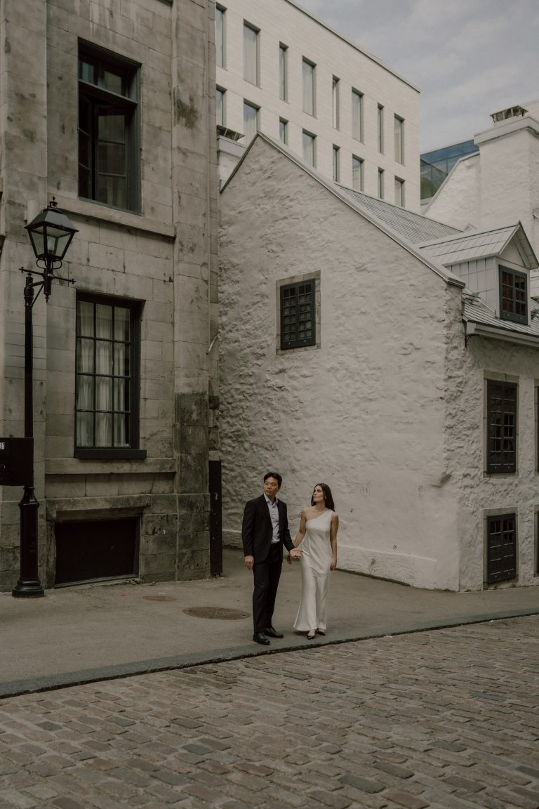 Couple élégant marchant main dans la main dans les rues pavées du Vieux-Montréal, lors d’une séance fiançailles au style urbain et romantique.