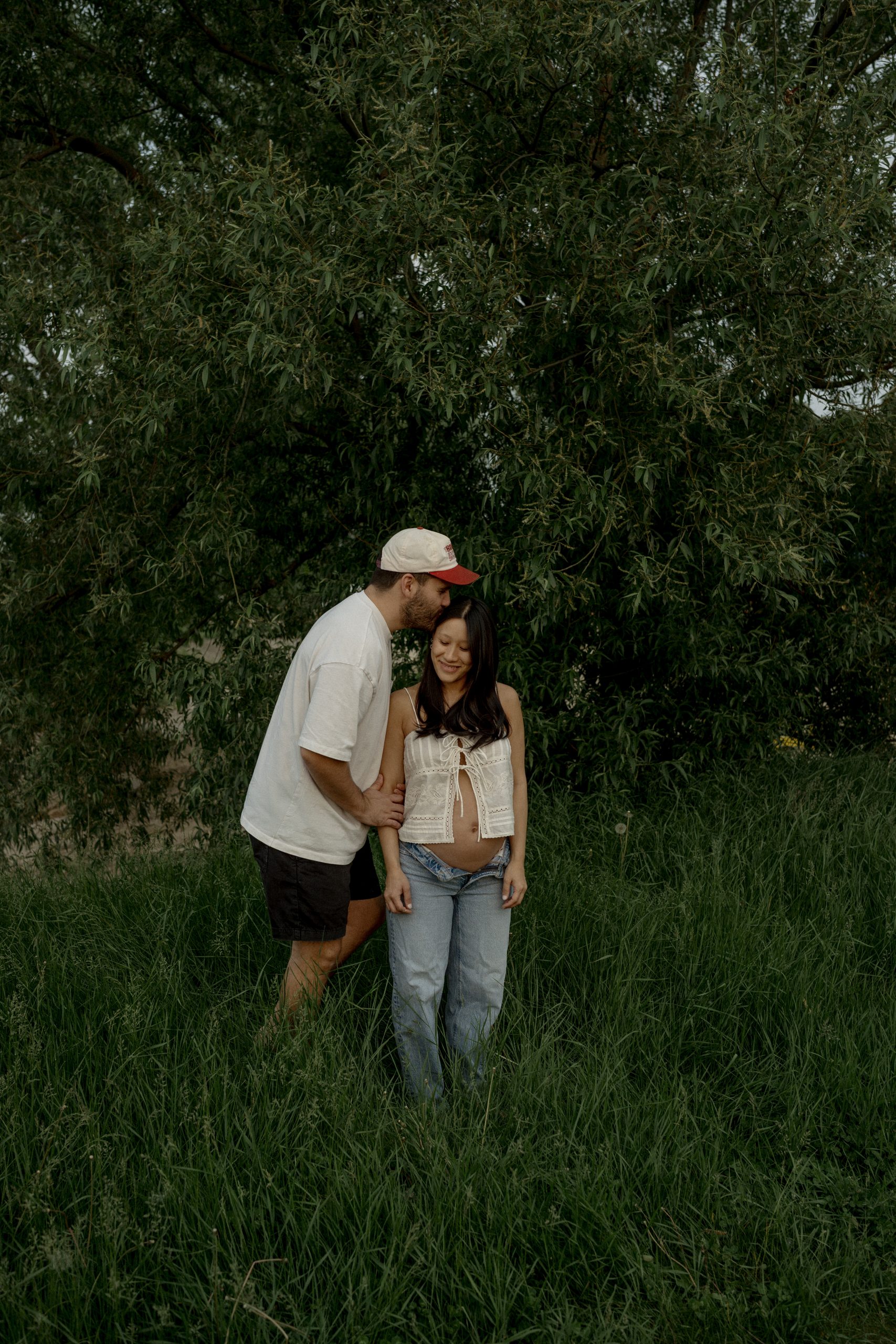 Couple en séance maternité lifestyle debout dans les herbes hautes, lumière naturelle douce et ambiance intime en plein air, photo professionnelle réalisée par une photographe lifestyle au Québec.