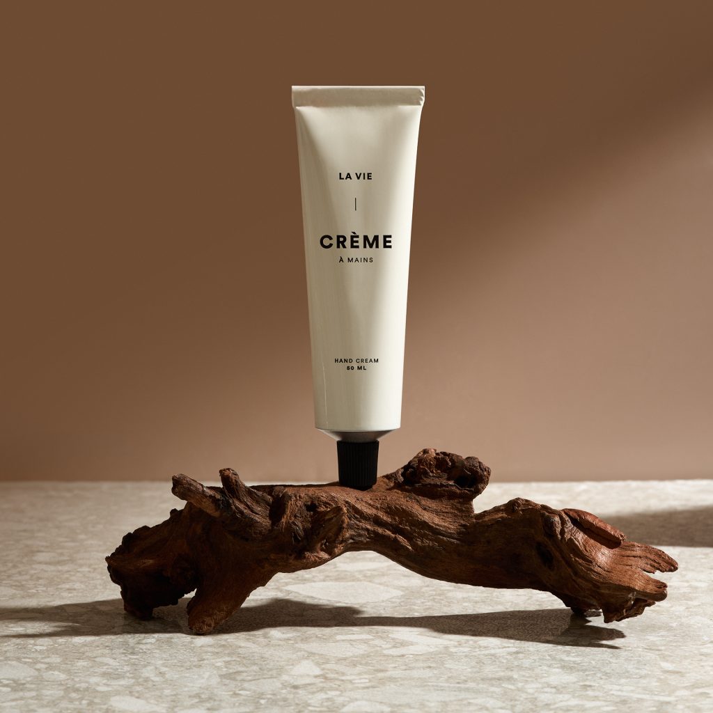 Tube de crème Atelier La Vie Apothicaire présenté sur une pièce de bois naturel dans une ambiance chaleureuse et organique. Photo commerciale professionnelle illustrant les ingrédients naturels, le bien-être et les soins corporels haut de gamme.