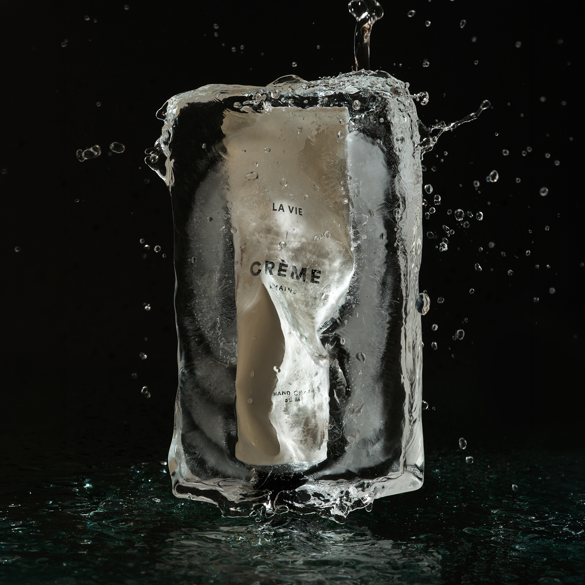Tube de crème Atelier La Vie Apothicaire emprisonné dans un bloc de glace traversé par des éclaboussures d’eau sur fond noir. Photographie de produit professionnelle mettant en valeur la fraîcheur, la pureté et l’efficacité des soins corporels naturels.