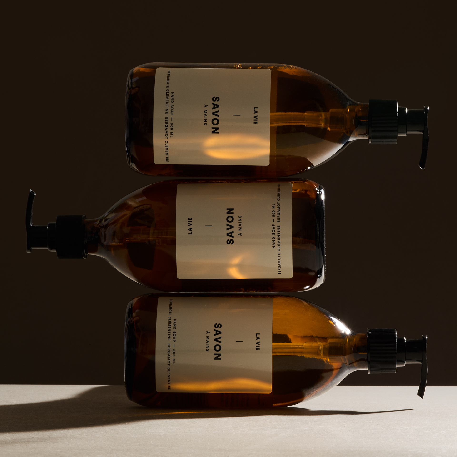 Flacons ambrés de savon Atelier La Vie Apothicaire empilés dans une composition minimaliste baignée d’une lumière chaude et enveloppante. Photo commerciale professionnelle réalisée pour une marque de soins naturels, idéale pour le commerce électronique et le branding.