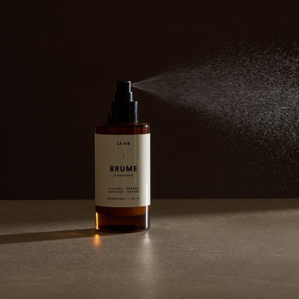 Flacon de brume Atelier La Vie Apothicaire diffusant un nuage de vaporisation dans une lumière dramatique sur fond sombre. Photographie de produit professionnelle mettant en valeur la texture aérienne du produit et l’univers raffiné de la marque.