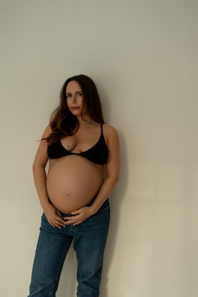 Femme enceinte appuyée contre un mur clair, portant un jean et un haut noir, mise en valeur par une lumière naturelle douce et une ambiance minimaliste. Photo professionnelle réalisée par une photographe de maternité au Québec, capturant la beauté de la grossesse dans un style intemporel et épuré.