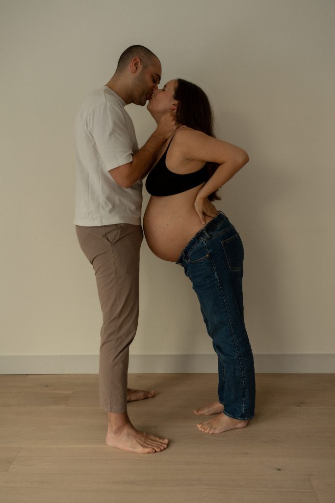 Future maman et son partenaire échangent un baiser dans un studio minimaliste aux tons neutres, créant une scène remplie de douceur et de complicité. Photo professionnelle réalisée par une photographe de grossesse au Québec, mettant en valeur l’amour et l’anticipation de cette étape de vie.