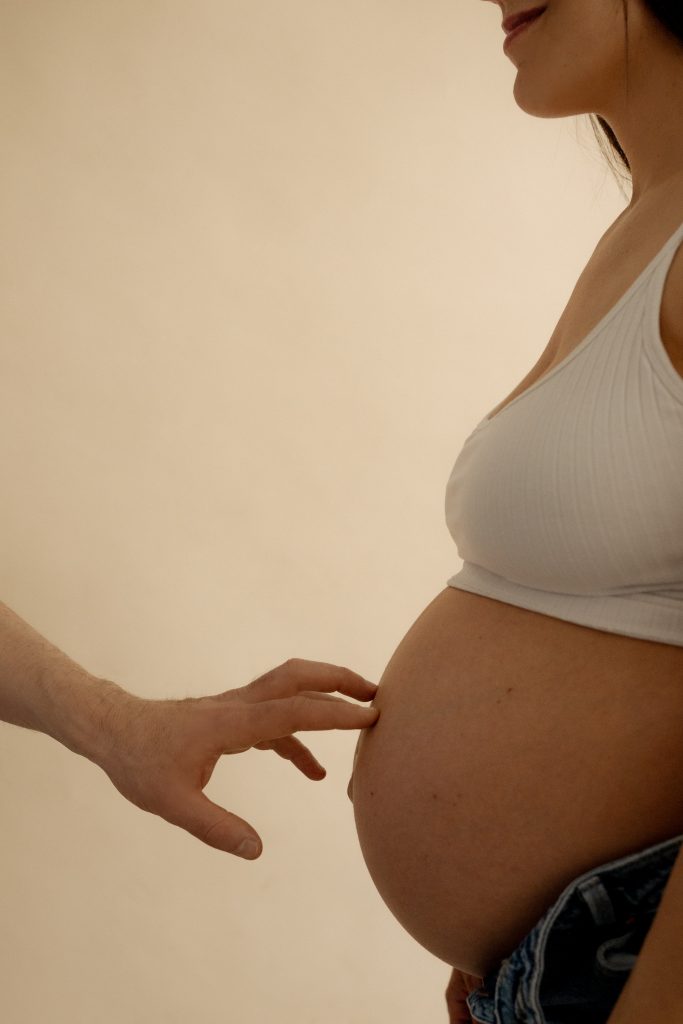 Gros plan du ventre arrondi d’une future maman alors qu’une main vient délicatement le toucher dans une lumière chaleureuse et enveloppante. Photo professionnelle réalisée par une photographe spécialisée en grossesse à Montréal, mettant en valeur les détails et l’émotion de cette période unique.