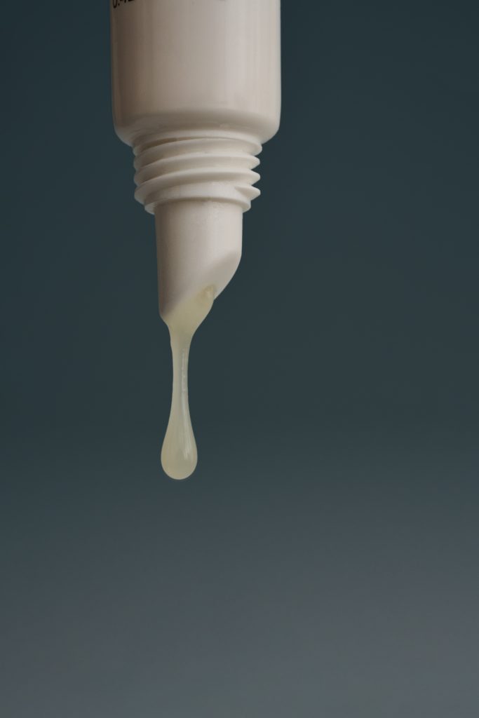 Gros plan d’un soin Nelly De Vuyst laissant couler une goutte de crème blanche sur un fond bleu foncé minimaliste. Photo professionnelle de cosmétique illustrant la texture du produit et la qualité des soins de la peau.
