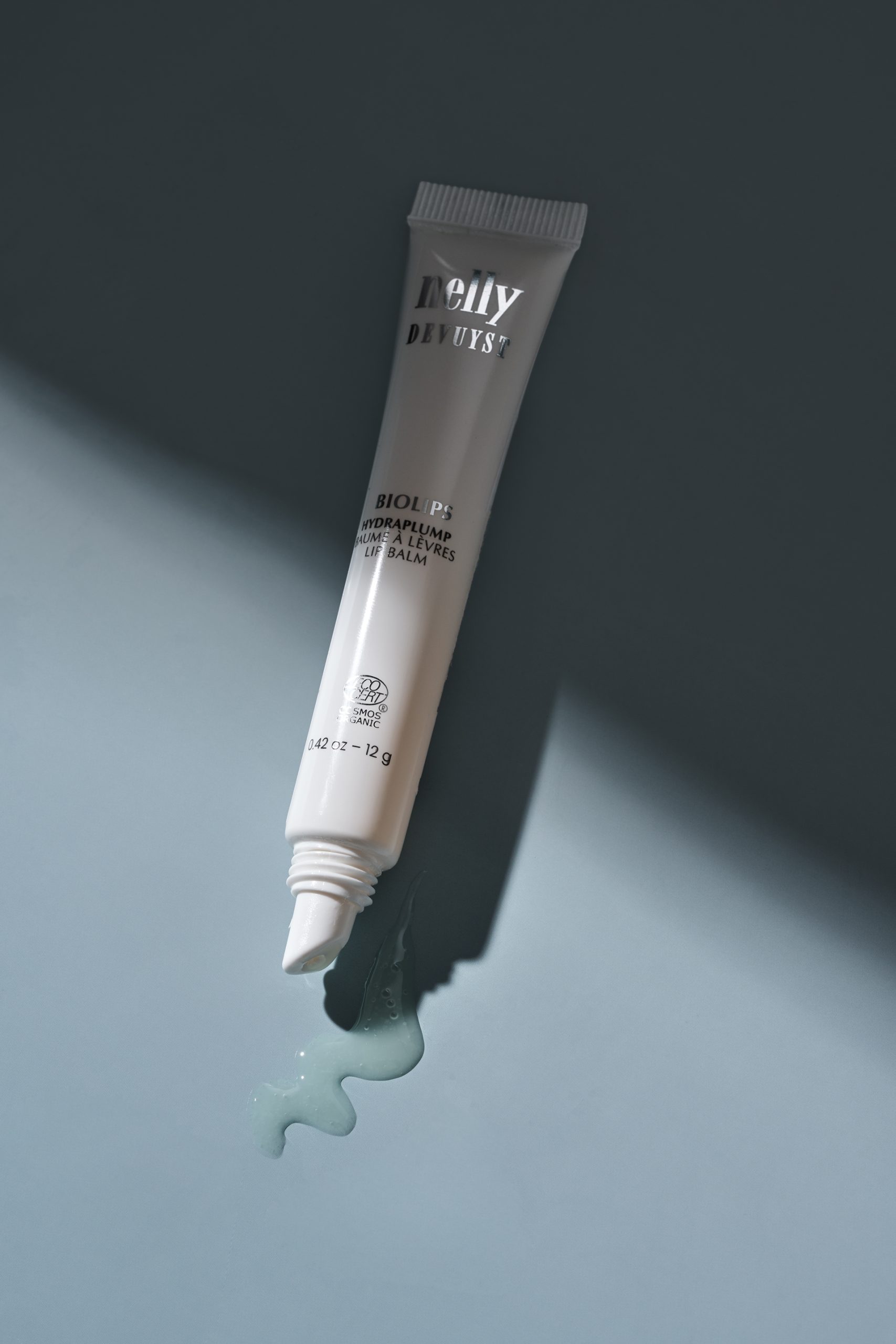 Tube de baume à lèvres BioTense de Nelly De Vuyst photographié sur une surface bleu profond avec une texture de produit visible et un éclairage dramatique. Photo professionnelle de cosmétique réalisée pour le commerce électronique, le branding et le marketing beauté.
