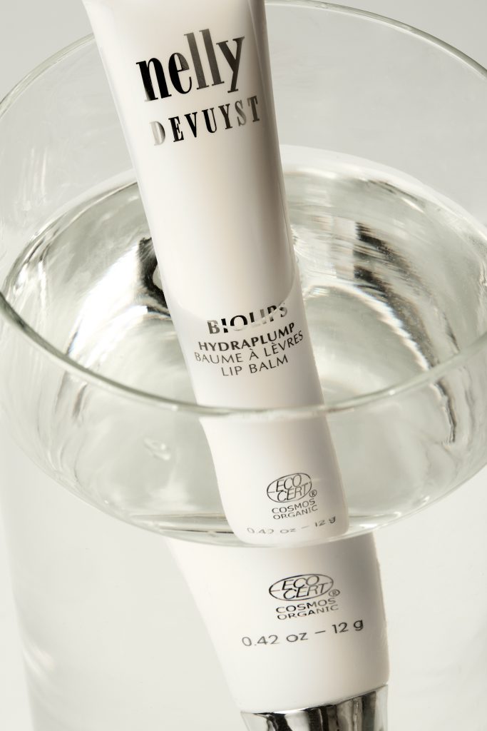 Tube de baume à lèvres BioTense de Nelly De Vuyst placé dans un verre d’eau transparent, créant des reflets lumineux et une ambiance fraîche et épurée. Photographie de produit professionnelle réalisée pour une marque de soins, idéale pour le commerce électronique et la publicité beauté.