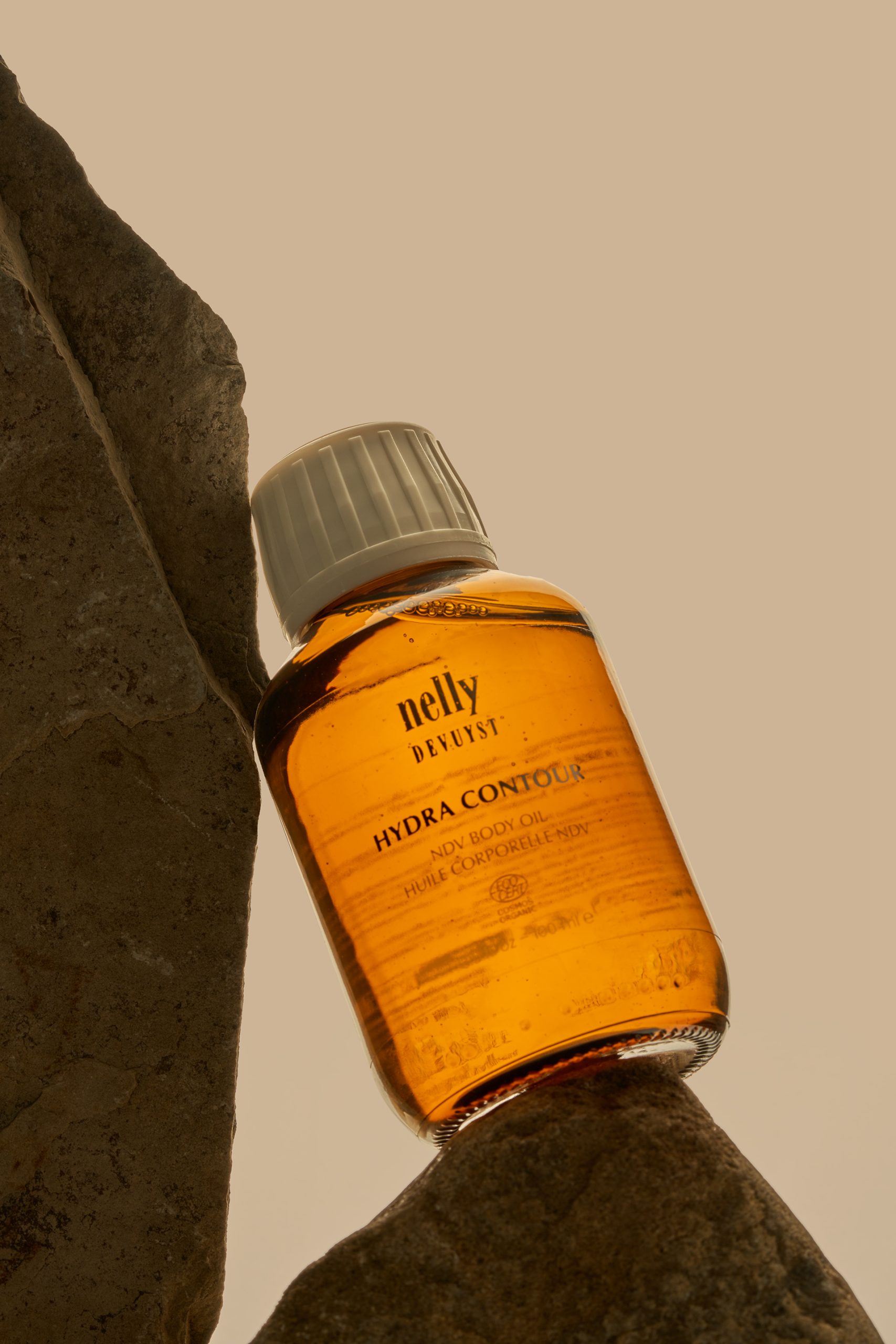 Flacon d’huile nettoyante Hydra Contour de Nelly De Vuyst mis en scène entre deux pierres naturelles dans une lumière chaude et minimaliste. Photographie de produit professionnelle réalisée pour une marque de soins de la peau, mettant en valeur la texture, les couleurs et l’univers haut de gamme du produit.