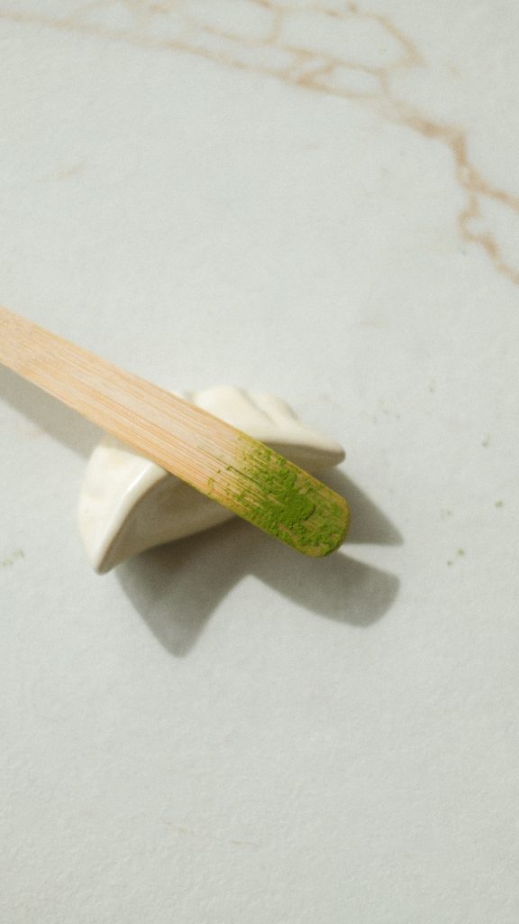 Cuillère traditionnelle en bambou déposée sur un porte-cuillère en céramique, recouverte d’une fine poudre de matcha vert vif. Photo professionnelle de produit réalisée pour le commerce électronique et la création de contenu autour du thé japonais.