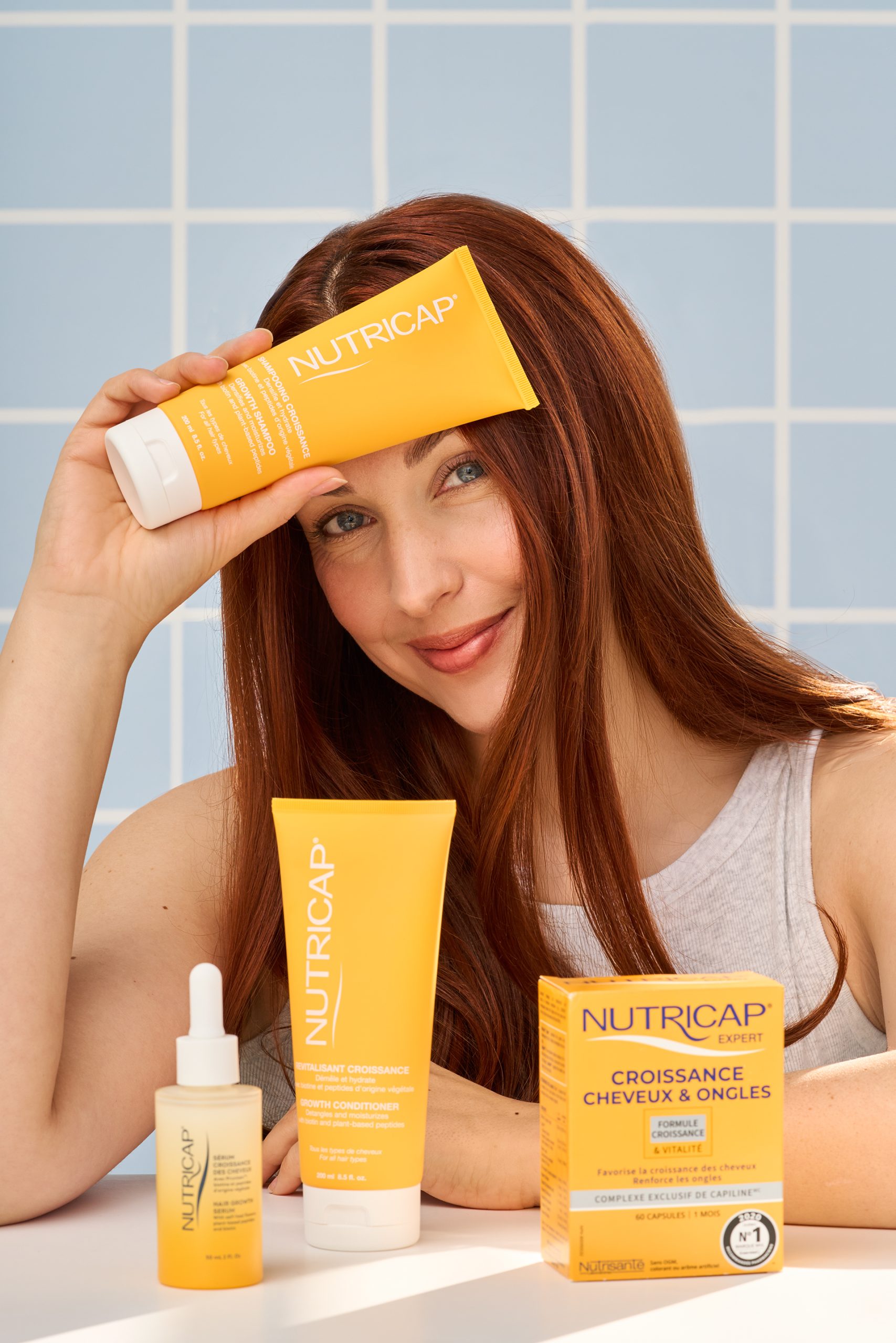 Femme aux longs cheveux roux présentant les produits Nutricap devant un fond de carreaux bleu pâle, dans une ambiance lumineuse et dynamique. Photographie commerciale professionnelle réalisée pour une marque de soins capillaires, mettant en valeur les produits et les résultats associés à la santé des cheveux.