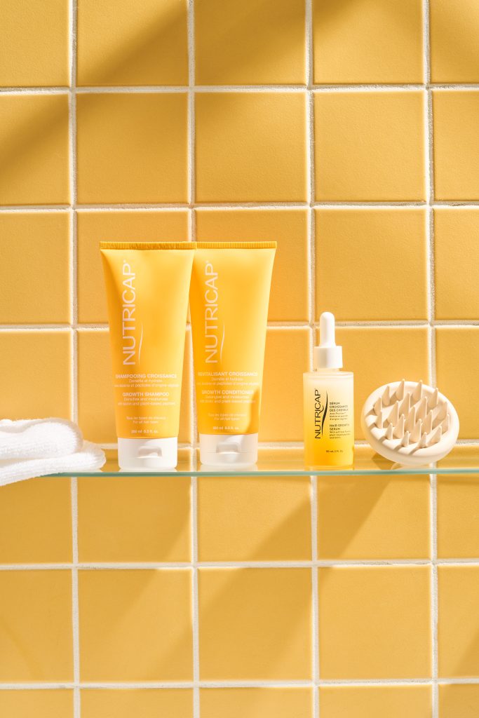 Produits Nutricap présentés sur une tablette devant un mur de carreaux jaunes lumineux, accompagnés d’accessoires de soins capillaires. Photographie commerciale professionnelle mettant en valeur l’identité visuelle de la marque et son univers de soins.