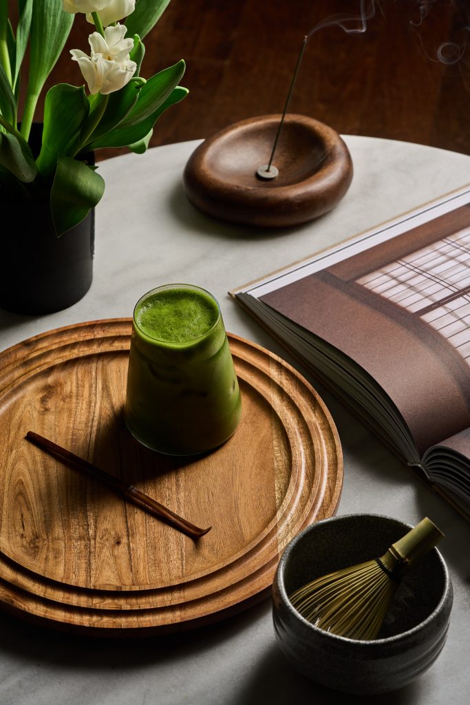 Mise en scène raffinée comprenant un matcha préparé, des accessoires traditionnels japonais, un livre ouvert et des fleurs blanches dans une ambiance zen et chaleureuse. Photographie commerciale réalisée pour Maison Minori, évoquant le bien-être, la pleine conscience et le rituel du thé.