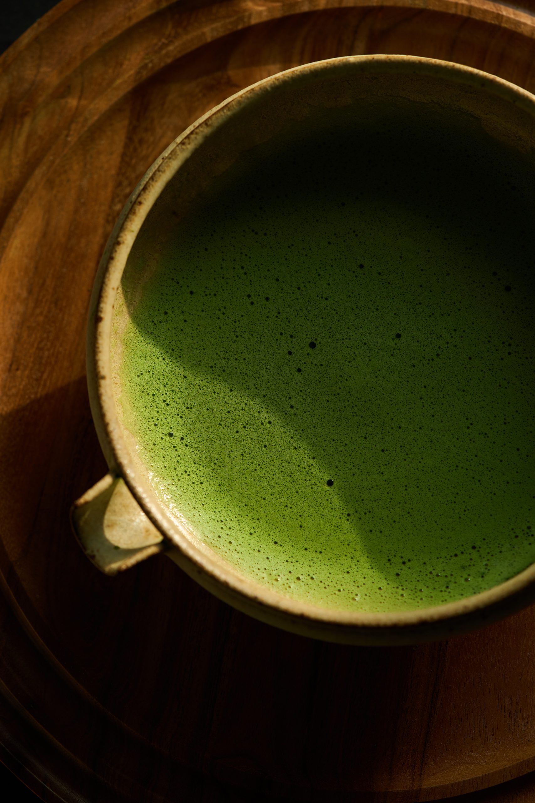 Bol de matcha cérémonial à la mousse délicate présenté sur un plateau en bois foncé, baigné par une lumière naturelle chaleureuse. Photographie de produit professionnelle réalisée pour Maison Minori, mettant en valeur le rituel japonais du thé et l’art de vivre slow living.