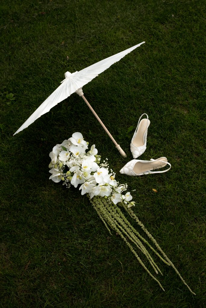 Photo de mariage présentant bouquet, chaussures de mariée et ombrelle blanche disposés sur l’herbe, photographie de mariage au Québec.