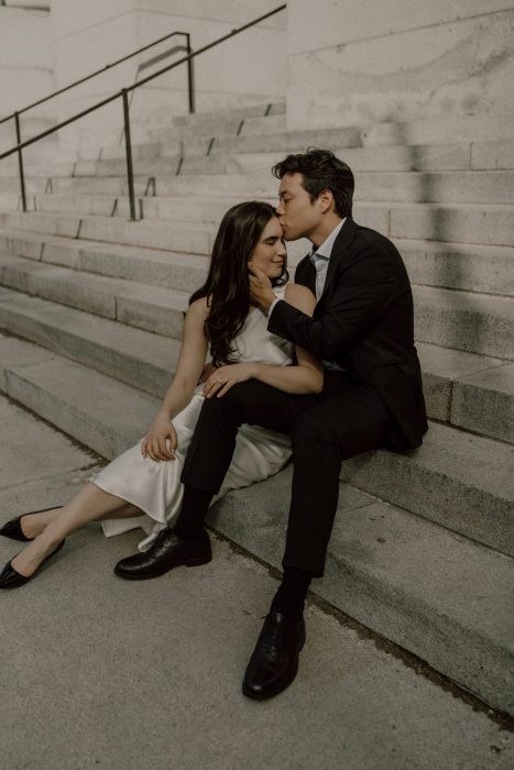 Moment tendre entre deux futurs mariés assis sur les marches en pierre d’un bâtiment, l’homme embrassant doucement le front de sa fiancée en robe blanche.