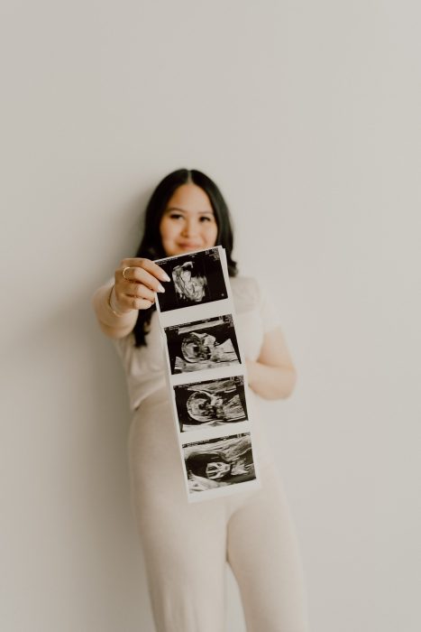 Femme enceinte tenant une série d'échographies en avant-plan, souriante, avec un fond clair et épuré.