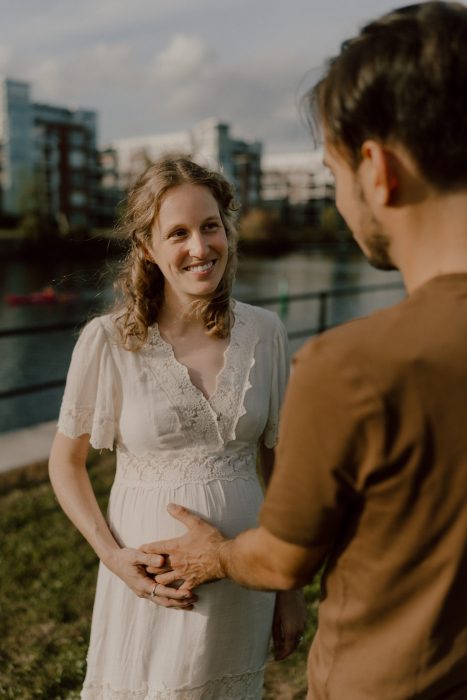 Femme enceinte souriante, en robe blanche, tenant son ventre pendant que son partenaire pose tendrement la main dessus, partageant un moment de complicité en extérieur.