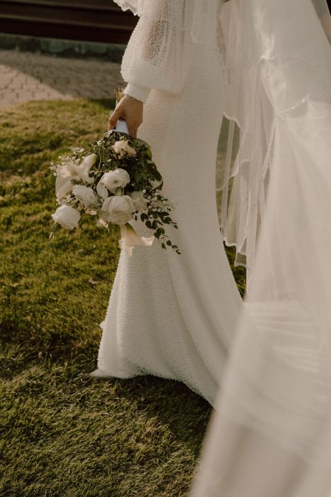 Détail du bouquet de la mariée tenu à la main avec robe et voile en mouvement sur fond naturel, photo de mariage élégante réalisée par une photographe professionnelle au Québec.