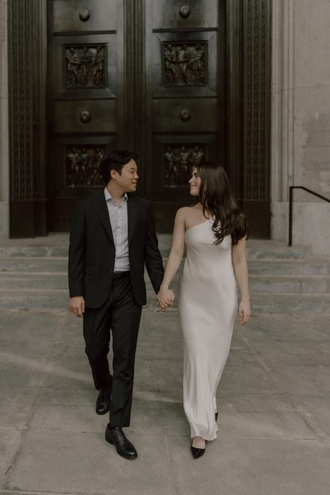 Couple élégant marchant main dans la main devant de grandes portes sculptées, échangeant un regard complice lors d’une séance fiançailles urbaine.