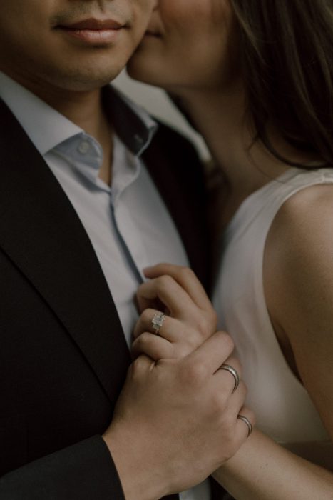 Gros plan intime d’un couple se tenant la main, mettant en valeur la bague de fiançailles scintillante et l’élégance de leur tenue.