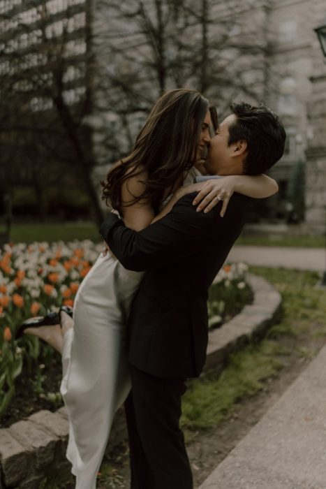 Couple de futurs mariés s’enlaçant avec joie dans un jardin fleuri, la mariée en robe blanche étant soulevée dans les bras de son partenaire en costume noir.