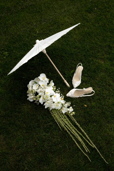 Photo de mariage présentant bouquet, chaussures de mariée et ombrelle blanche disposés sur l’herbe, photographie de mariage au Québec.