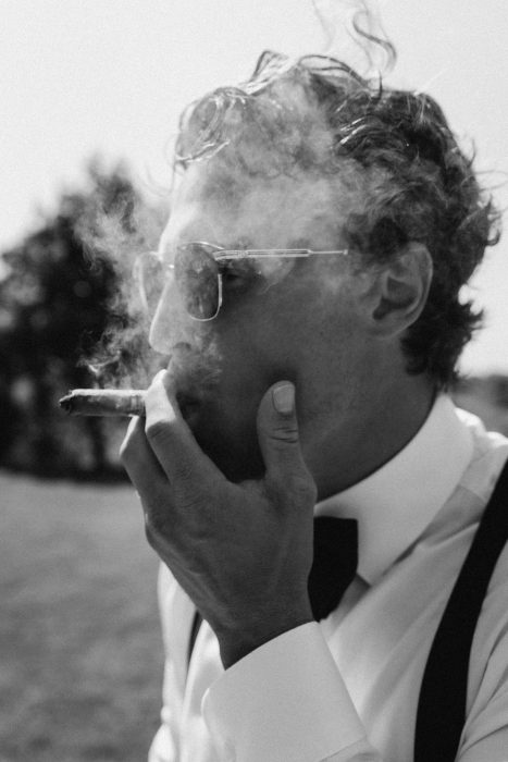 Portrait du marié en noir et blanc fumant un cigare, portant bretelles et nœud papillon à l’extérieur, photo de mariage artistique réalisée par une photographe professionnelle au Québec.
