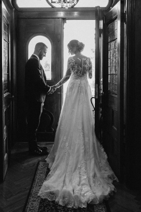 Photo en noir et blanc d'un couple de mariés tenant leurs mains dans l'entrée d'une maison ancienne, la mariée de dos en robe longue.