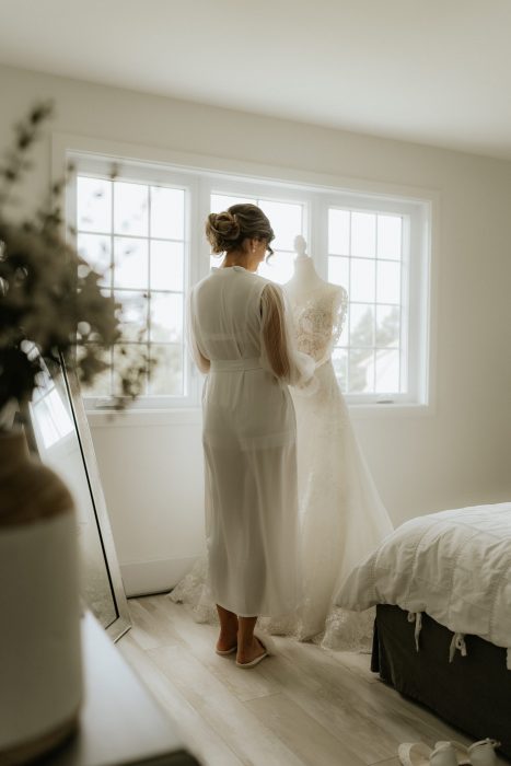 Mariée debout devant sa robe de mariée dans une chambre lumineuse, regardant sa tenue avec émotion.